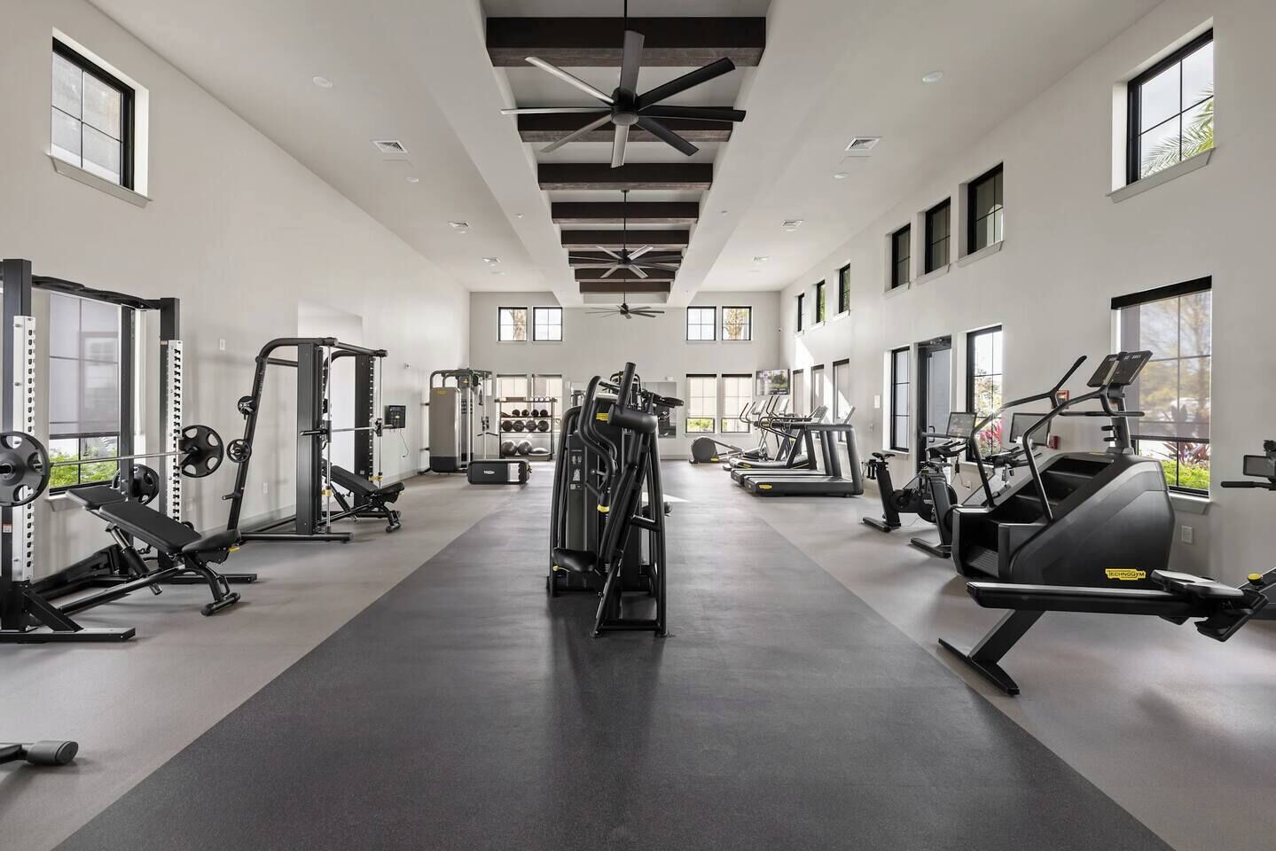 Ein geräumiger Fitnessraum mit unterschiedlichen Trainingsgeräten, großen Fenstern und einer hohen Decke.