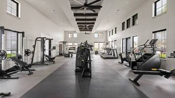 Ein geräumiger Fitnessraum mit unterschiedlichen Trainingsgeräten, großen Fenstern und einer hohen Decke.