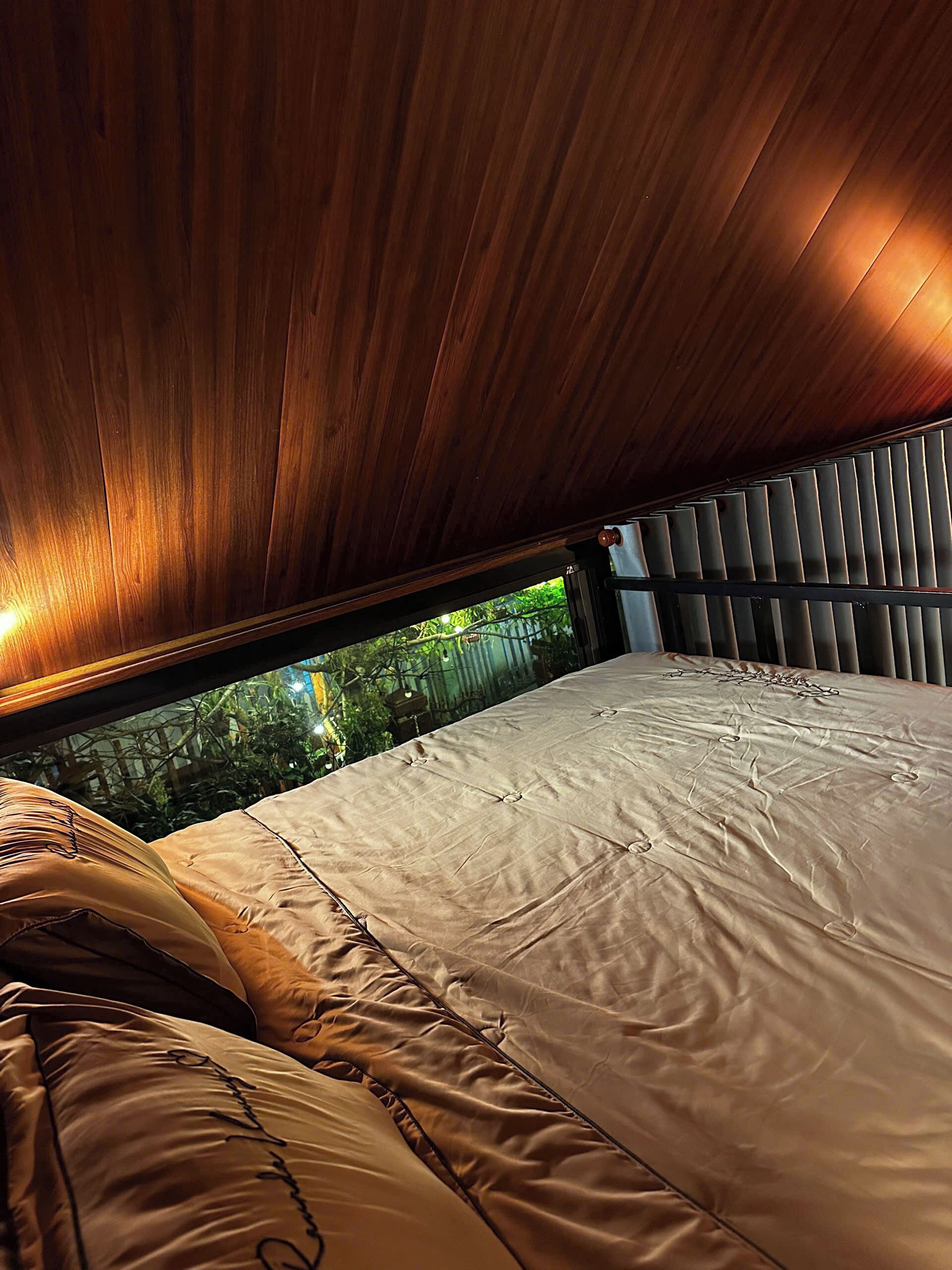A bedroom with a wooden ceiling, a bed with a beige bedspread, and a view of greenery outside.