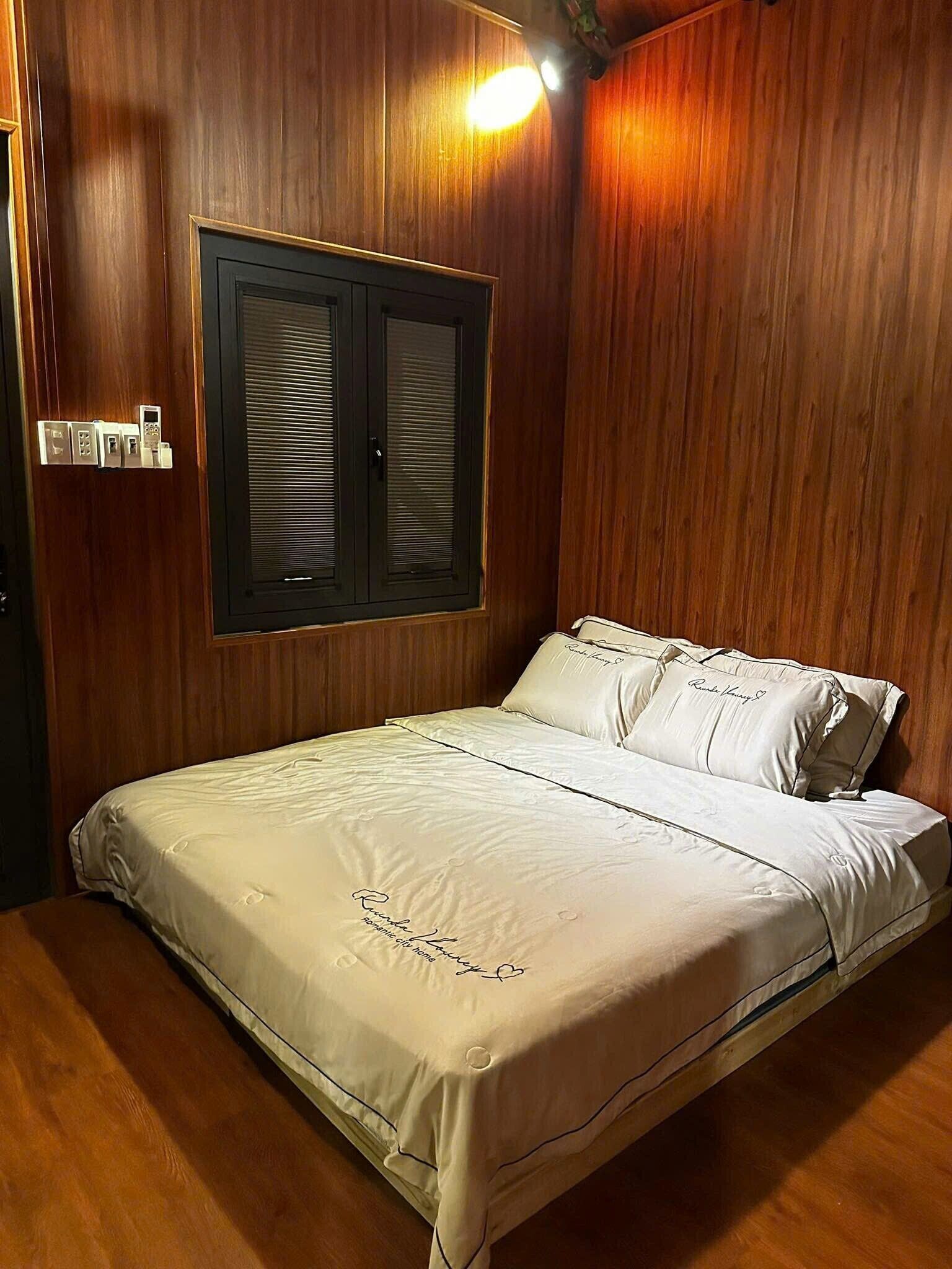 A bedroom with a wooden wall, a bed with white bedding, a window with shutters, and a wall-mounted light fixture.