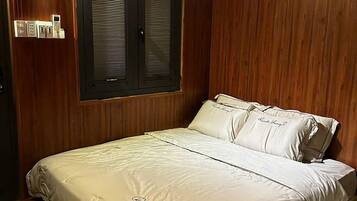 A bedroom with a wooden wall, a bed with white bedding, a window with shutters, and a wall-mounted light fixture.