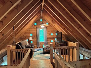 Et rum på loftet med skrå vægge, træbjælker, en loftsvifte, et vindue med gardiner, en sofa, en lampe og et lille bord.