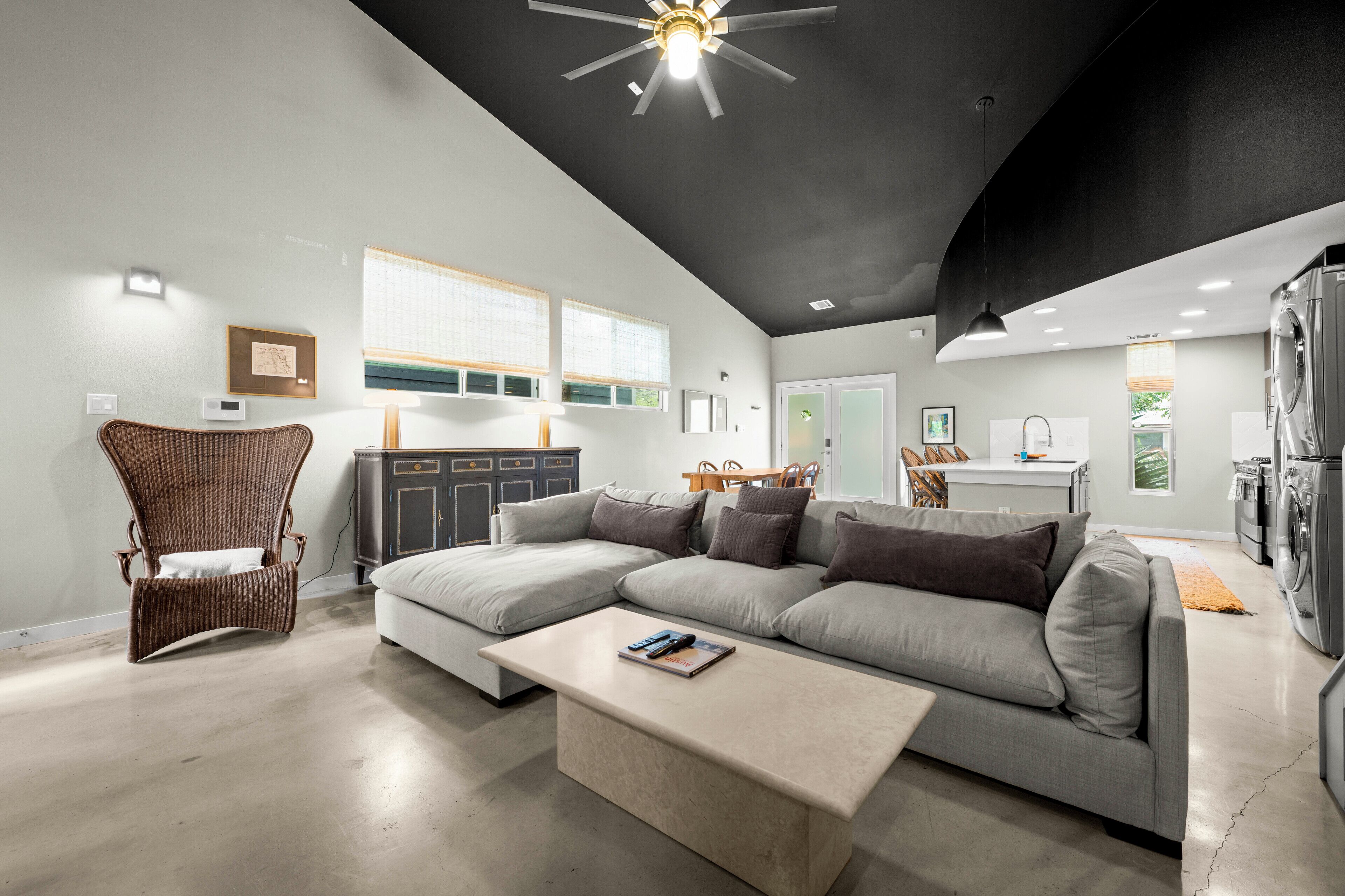 A modern living room with a sectional sofa, a coffee table, a wicker chair, and a kitchen area with appliances.