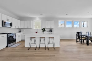 A modern kitchen with white cabinets, a central island with stools, a dining area with a table and chairs, and large windows.