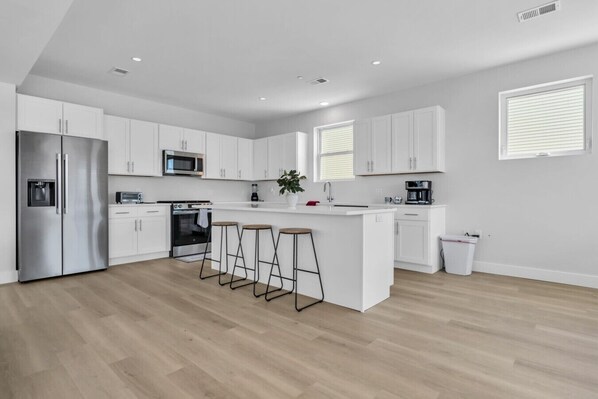 A modern kitchen with white cabinetry, a stainless steel refrigerator, a microwave, a coffee machine, and a small island with stools.
