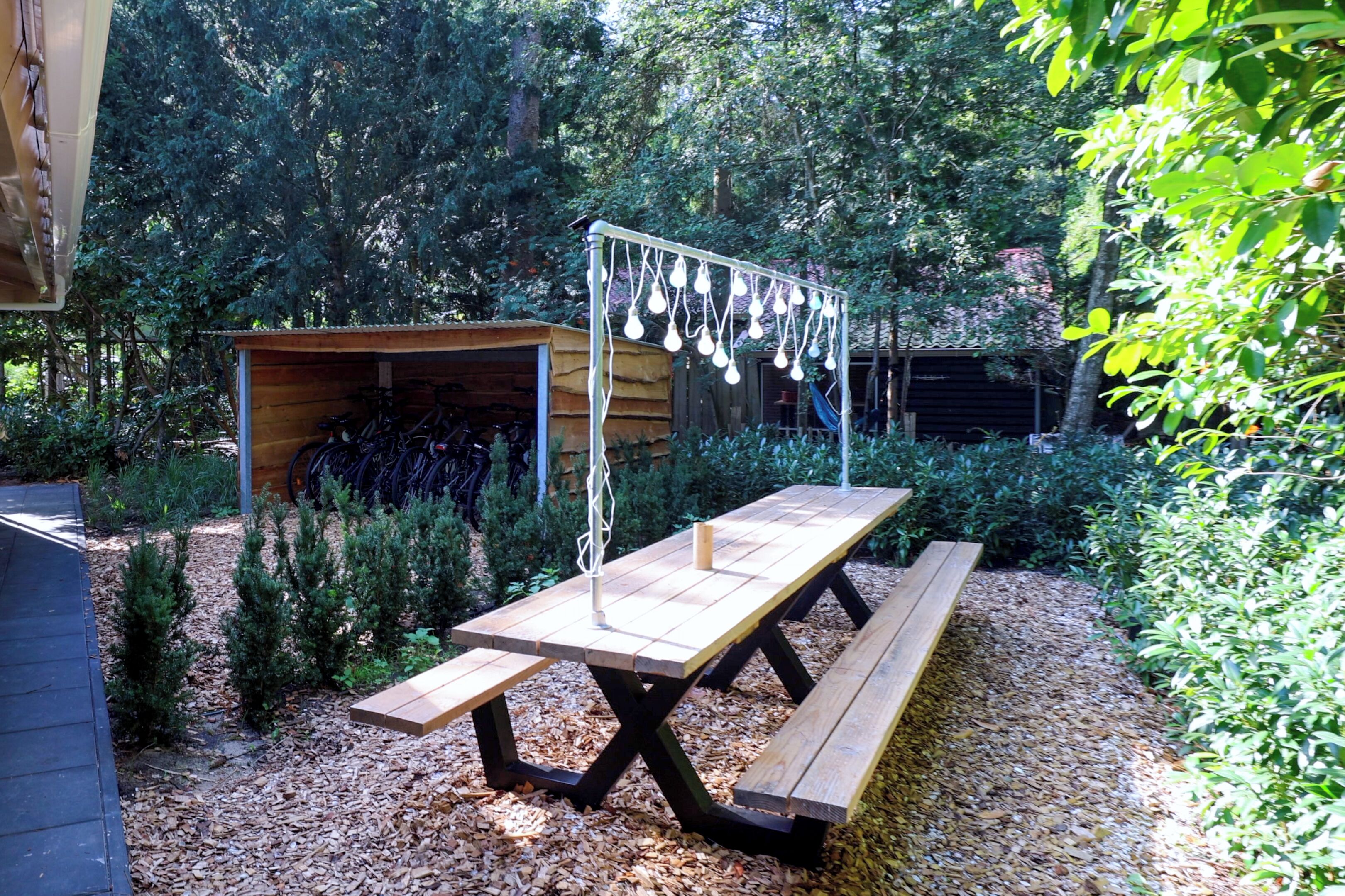 A wooden picnic table with benches, surrounded by a garden with various plants and a shed in the background.