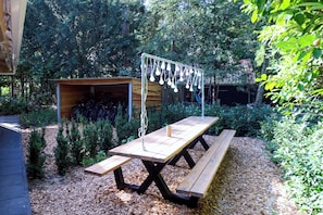 A wooden picnic table with benches, surrounded by a garden with various plants and a shed in the background.