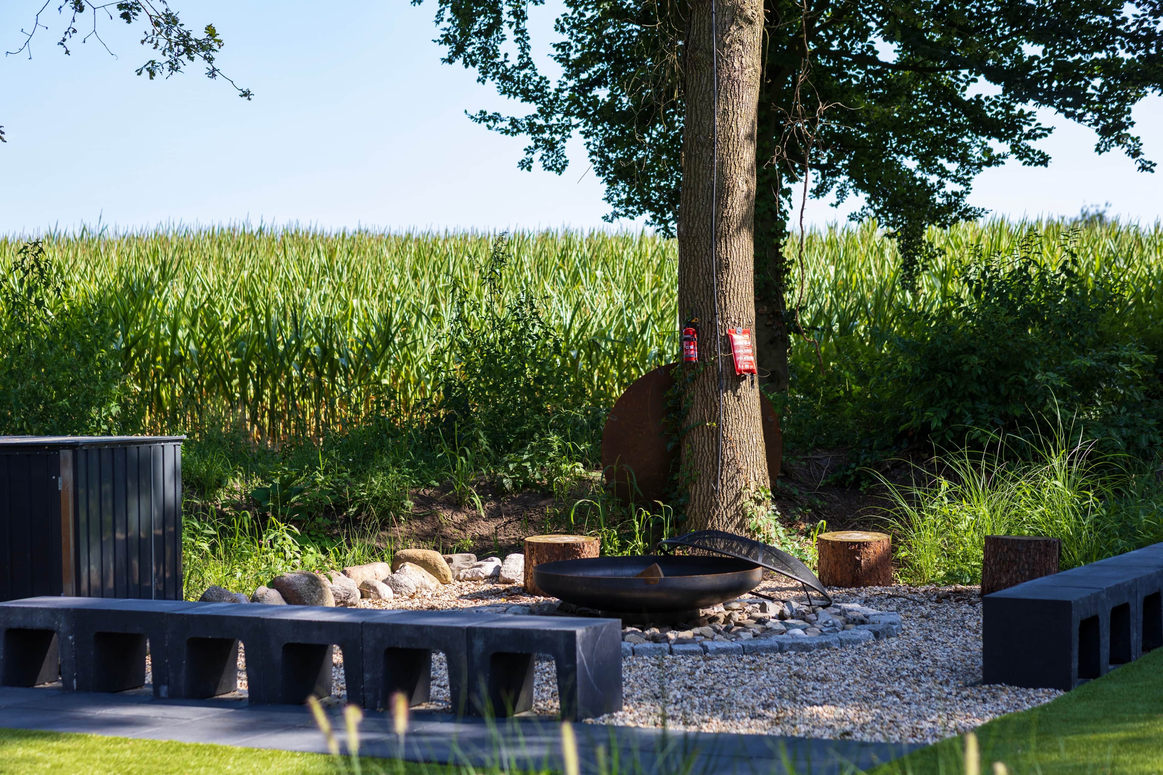 A rustic outdoor seating area with a fire pit, surrounded by a gravel path and wooden benches, set against a backdrop of a cornfield.