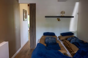 Habitació amb dos llits, cada un amb una manta blava i un coixí, una petita estant amb un objecte decoratiu i una porta que dona a una altra habitació.