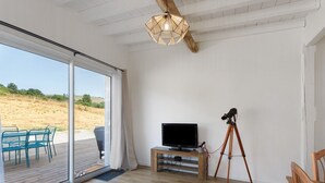Habitación con televisión sobre un pedestal de madera, un trípode y un amplio ventanal corredera que da a un área exterior con sillas.