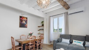 Un salón moderno con comedor, un sofá gris con almohadones, una mesa de comedor de madera con sillas, una lámpara colgante y una ventana con cortinas.