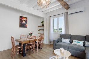 A modern living room with a dining area, a grey sofa with pillows, a wooden dining table with chairs, a hanging light fixture, and a window with curtains.