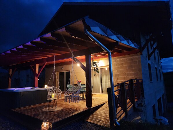 A covered outdoor seating area with chairs and a table, a hot tub, and a wooden deck.