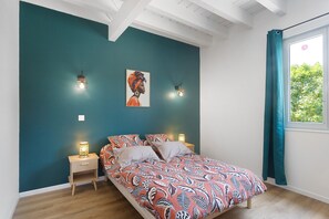 A bedroom with teal walls, a bed with patterned bedding, two bedside lamps, a framed artwork, and a window with curtains.