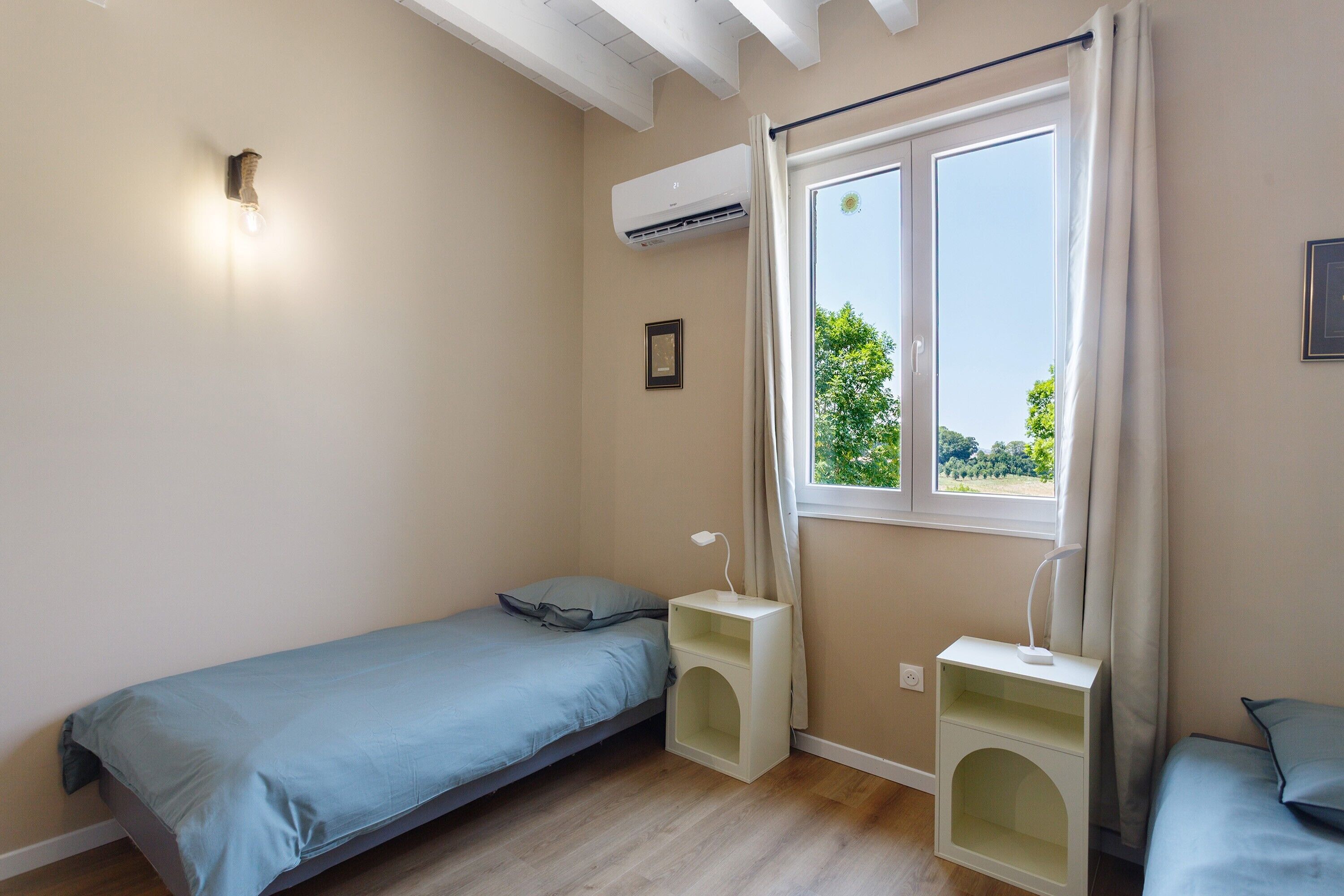 Ein kleiner, gut beleuchteter Raum mit einem Einzelbett, einem Nachttisch, einem Fenster mit Vorhängen und einer Klimaanlage.