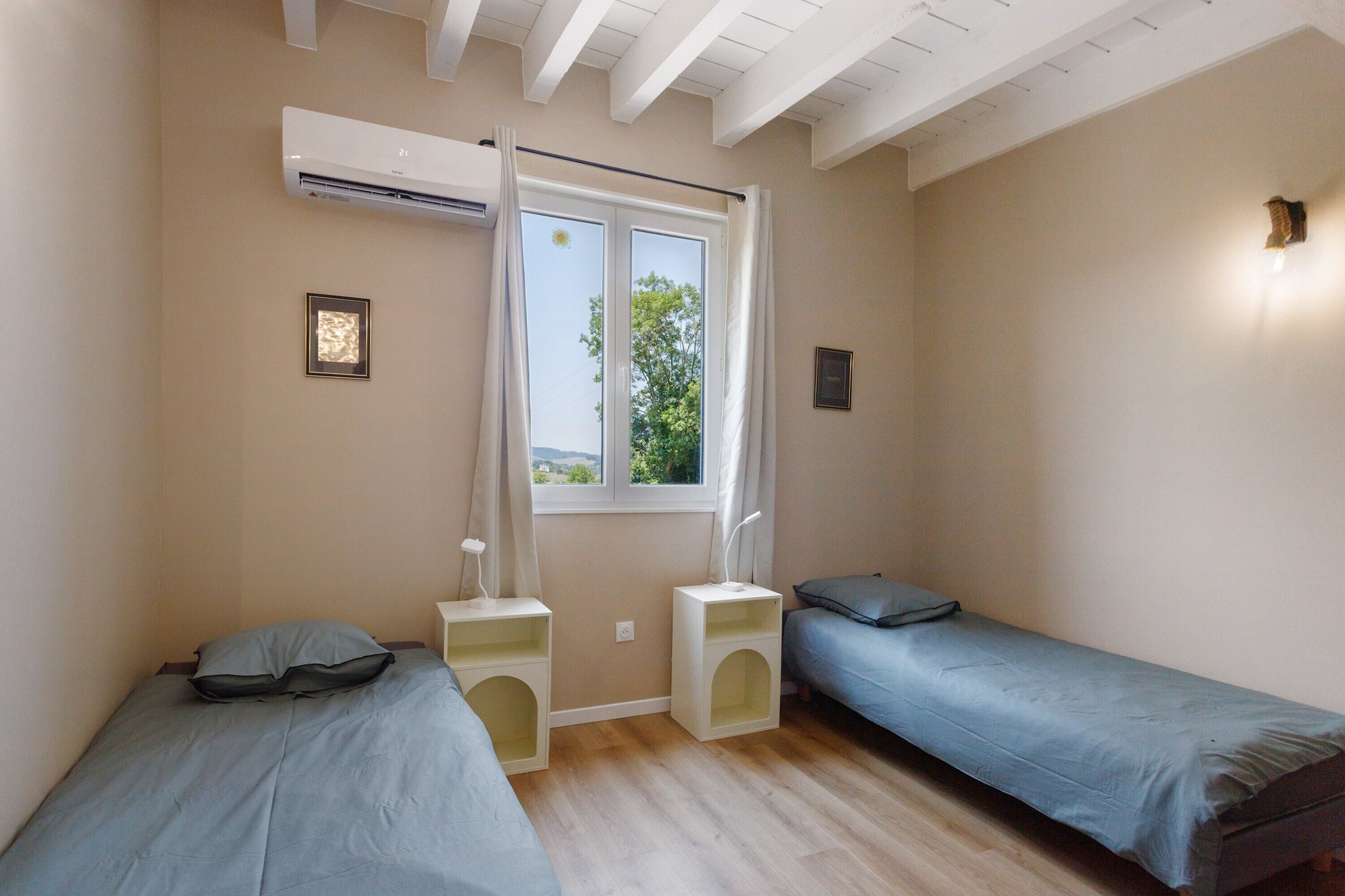 Une chambre avec deux lits simples, une fenêtre avec des rideaux, un climatiseur fixé au mur et une petite étagère avec une lampe.