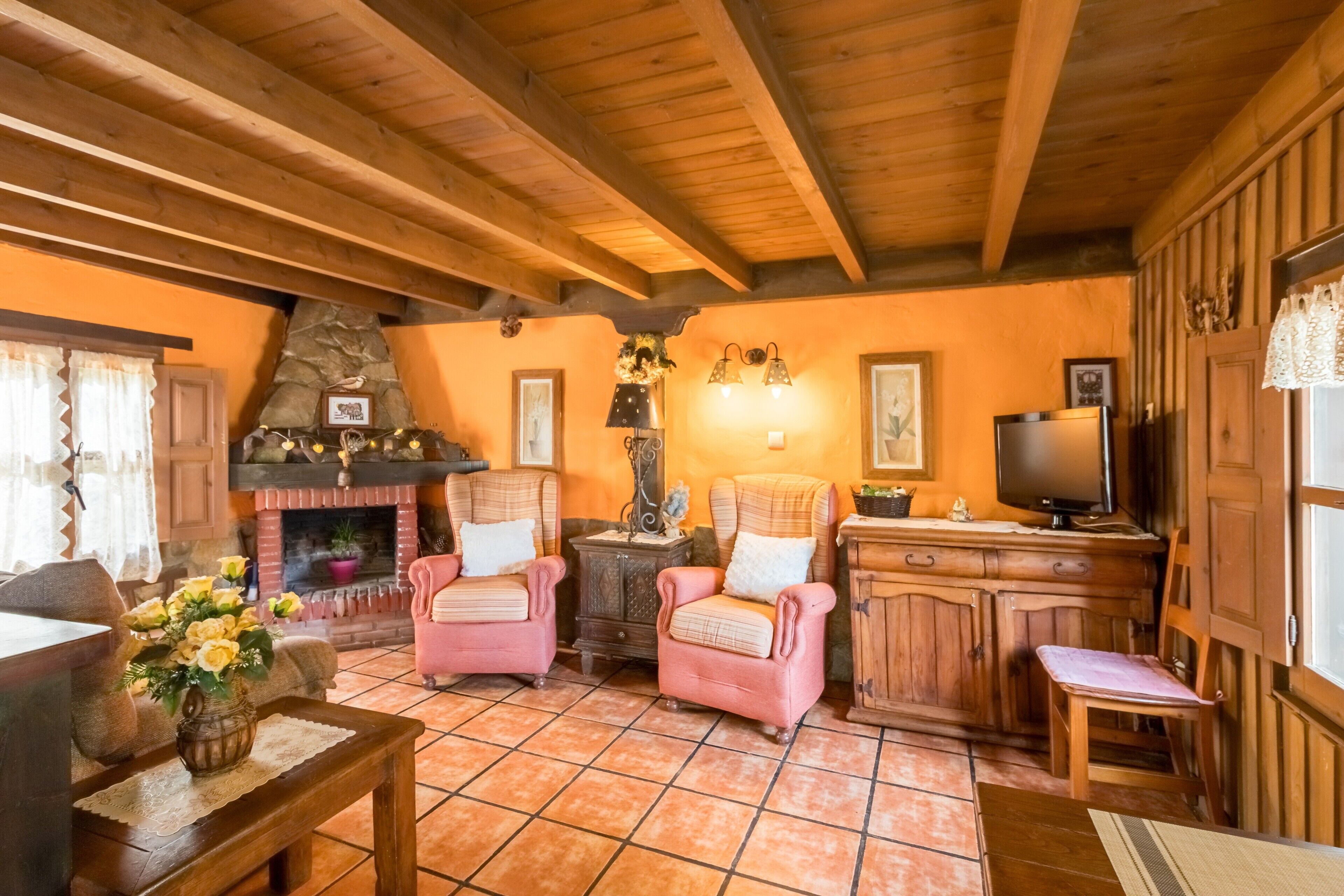 Een gezellige woonkamer met houten balken, een open haard, roze fauteuils, een televisie en een keukenhoek.
