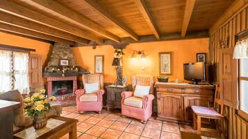 Een gezellige woonkamer met houten balken, een open haard, roze fauteuils, een televisie en een keukenhoek.