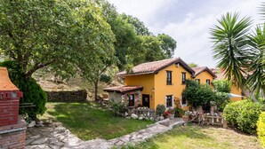 A yellow building with a stone pathway, surrounded by greenery and a stone wall.