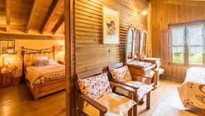 A wooden cabin interior with a bed, two wooden chairs, a nightstand, a mirror, and a window with curtains.