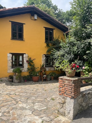 Un edifici de color groc amb marcaments de pedra a les finestres, un pati empedrat i plantes en testos.