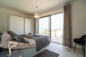 Ein Schlafzimmer mit einem Bett, einem Sessel, einem Nachttisch, einem großen Fenster und Blick ins Freie.