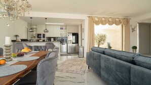 Sala de estar moderna com sofá, mesa de jantar, lustre e cozinha ao fundo.