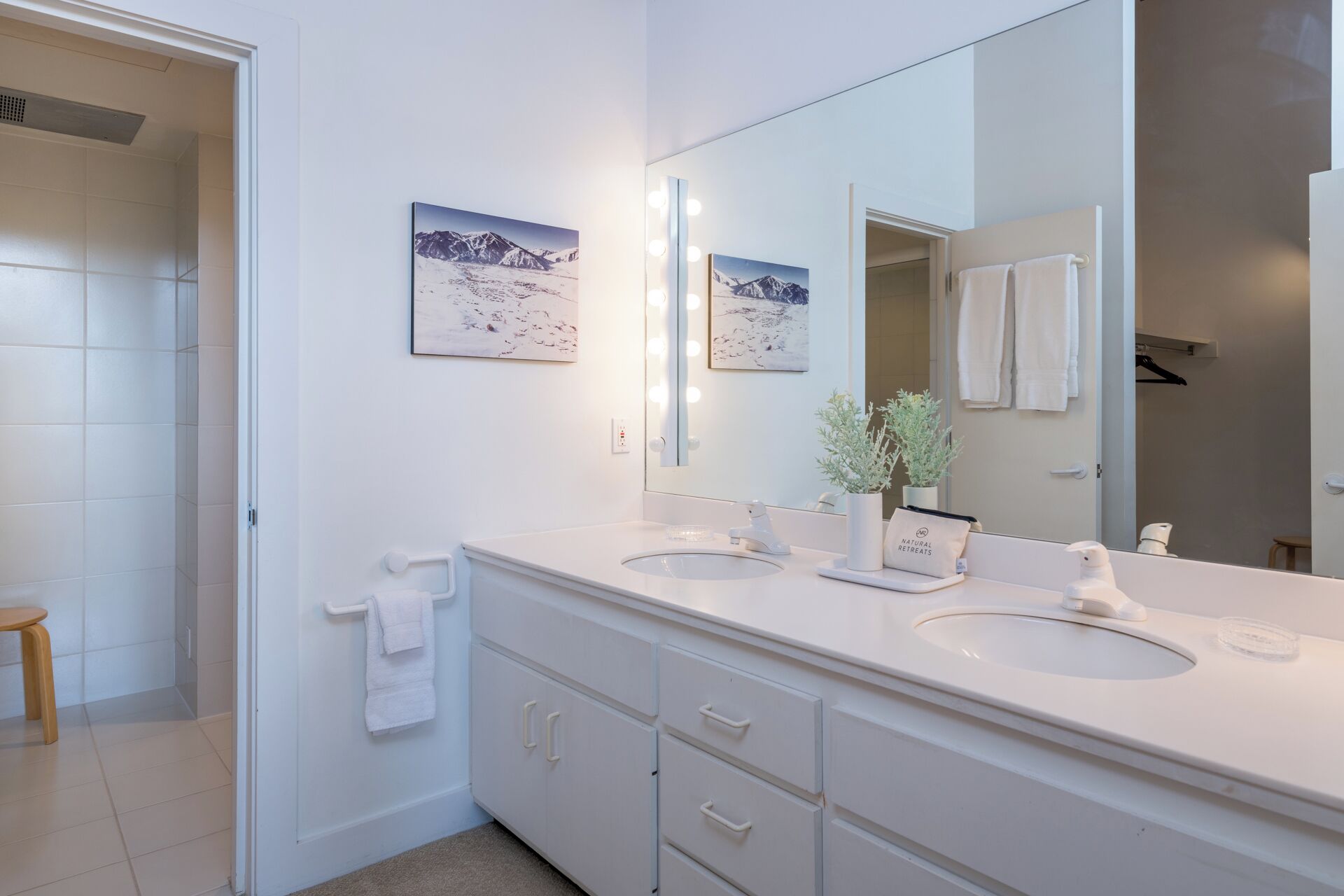 A bathroom with two sinks, a large mirror with lights, and two framed pictures on the wall.