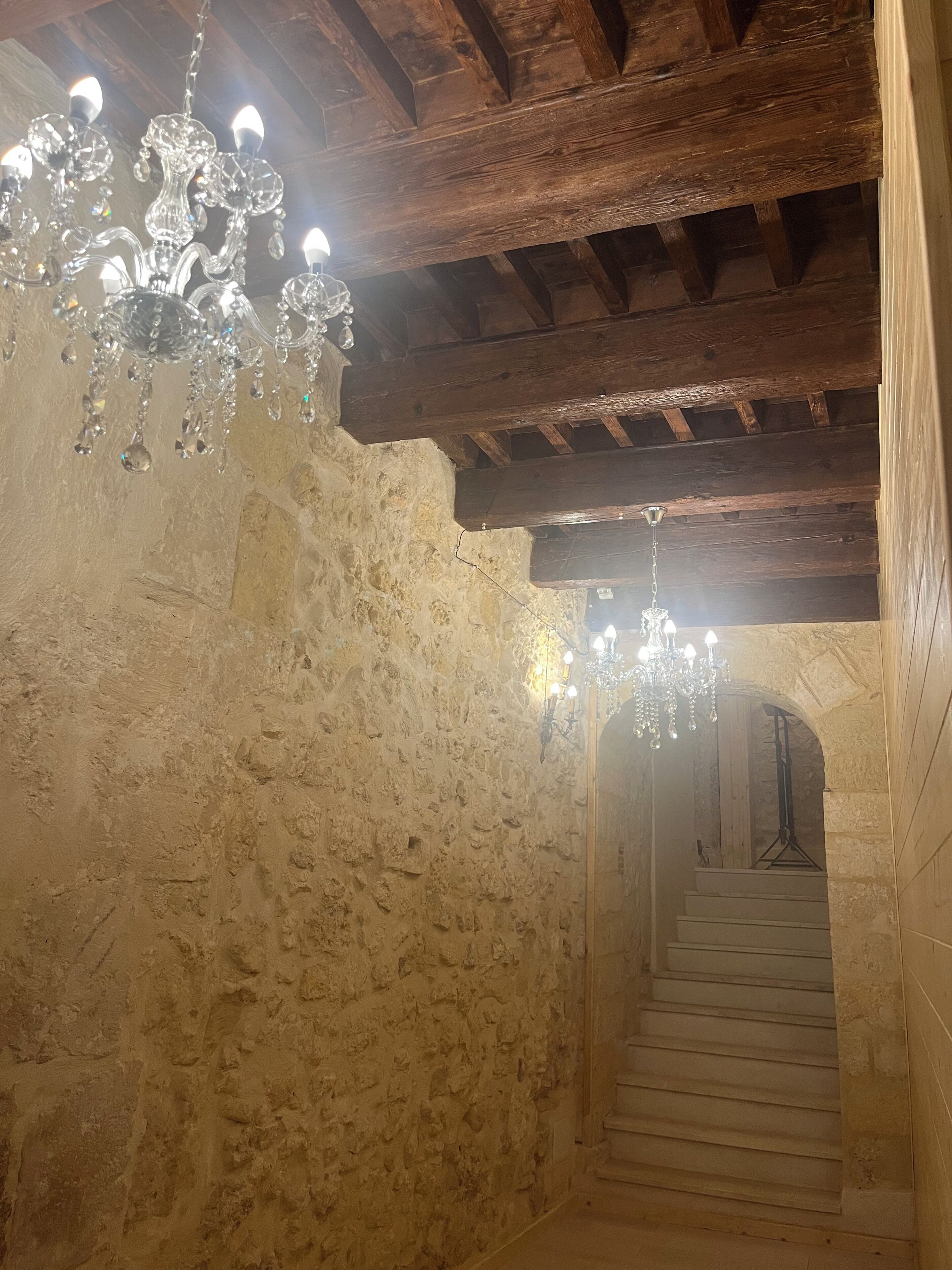 A stone-walled hallway with a wooden ceiling and chandeliers.