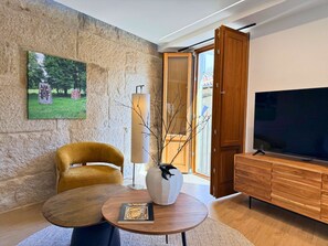 A modern living room with a stone wall, a yellow armchair, a round wooden table, a white vase, a television, and a wooden cabinet. - YourHouse O Rexo family apartment (Allariz)