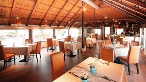 Restaurante com várias mesas e cadeiras, teto de madeira e vista para o exterior.