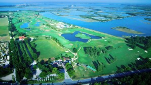 Vista aérea de um campo de golfe com vários corpos d'água, campos verdes e uma área residencial com casas e uma estrada.