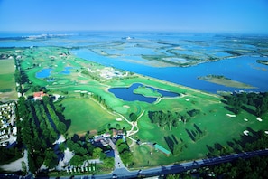 Vista aérea de un campo de golf con varios cuerpos de agua, campos verdes y una zona residencial con casas y una carretera.