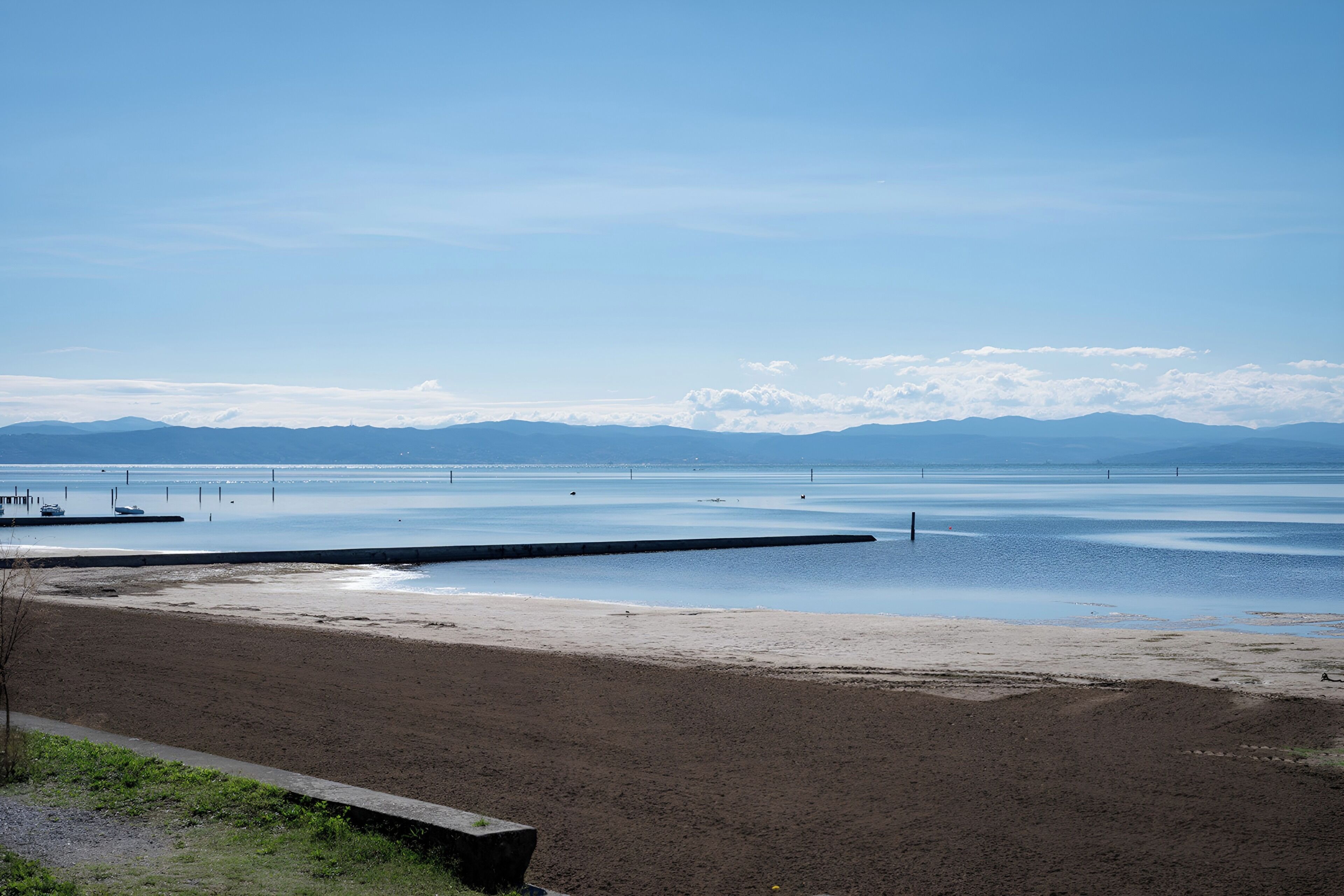 Een strand met een houten pier die zich uitstrekt over kalm water, een heldere lucht en verre bergen.