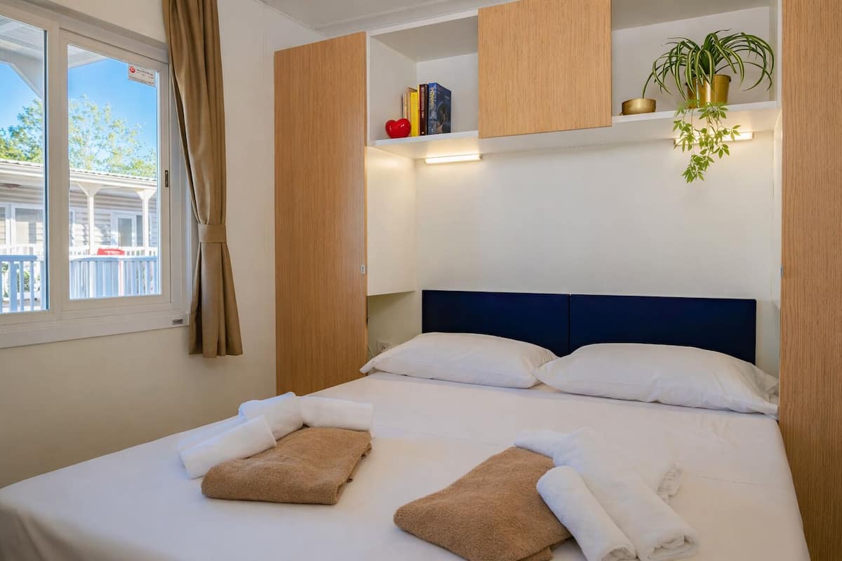 Habitación de hotel con cama, toallas, ventana, cortina, estante con libros y una planta, y vistas al balcón.