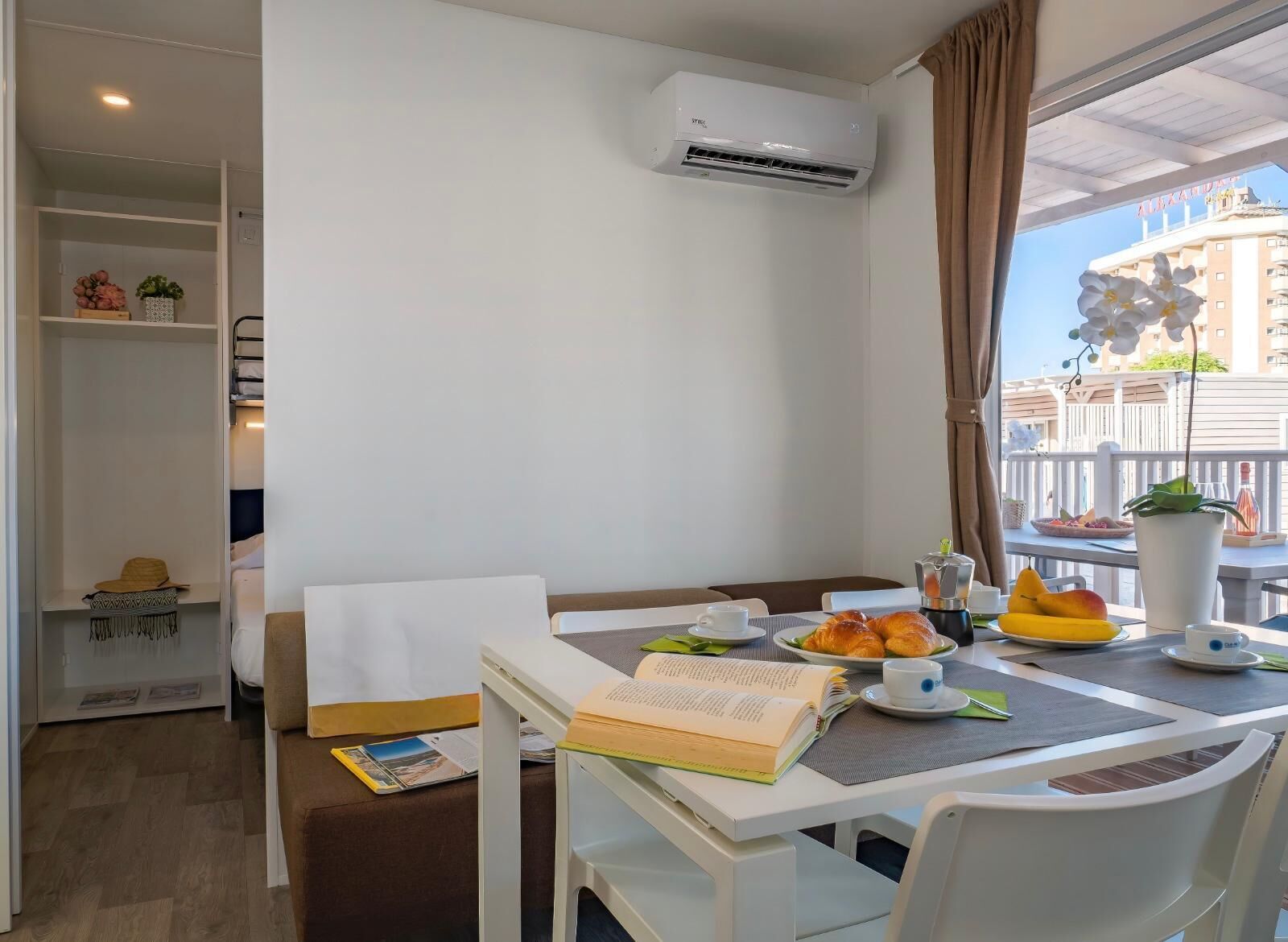 A hotel room with a dining table set with breakfast, a bookshelf, and a view of a building with balconies.