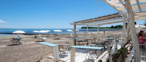 A beachside restaurant with outdoor seating, umbrellas, and a view of the sea.