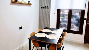 A dining area with a black table and wooden chairs, a small shelf with decorative items, a window with a view, and a door with a metal screen. - New!!! Loft Córdoba Home (Córdoba)