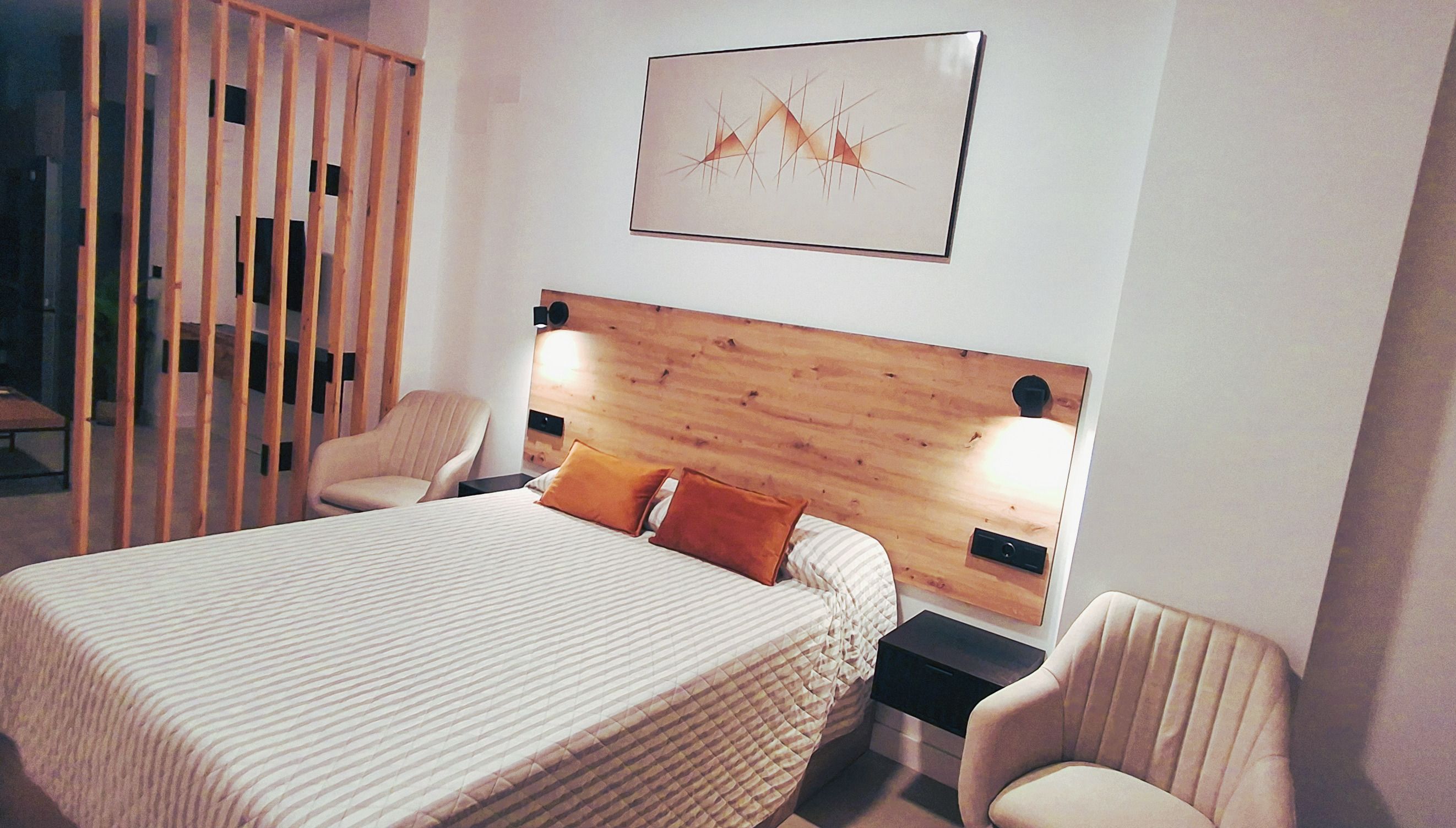 Una habitación de hotel con cama, dos sillas, una mesita de noche y cabecera de madera.