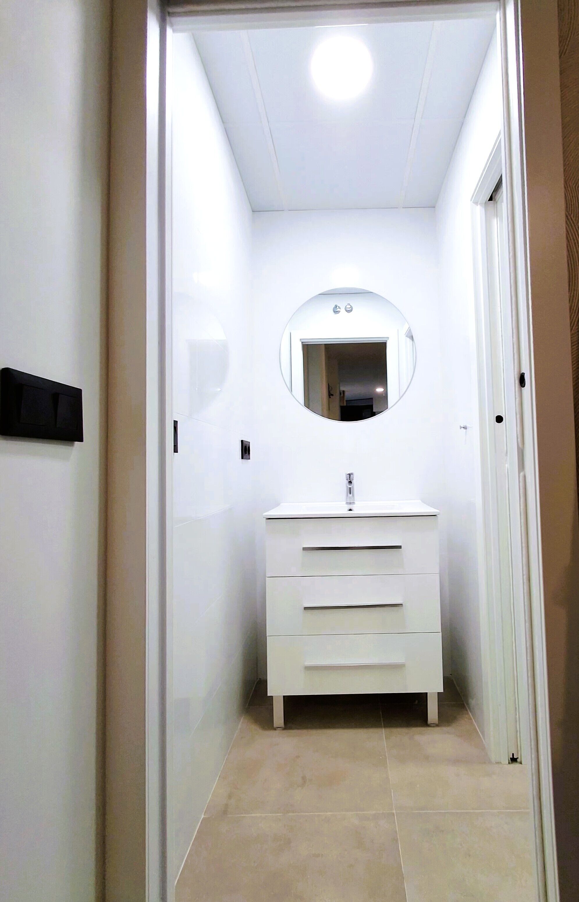 Un estrecho pasillo que conduce a un baño con un lavamanos blanco, un espejo y una puerta.