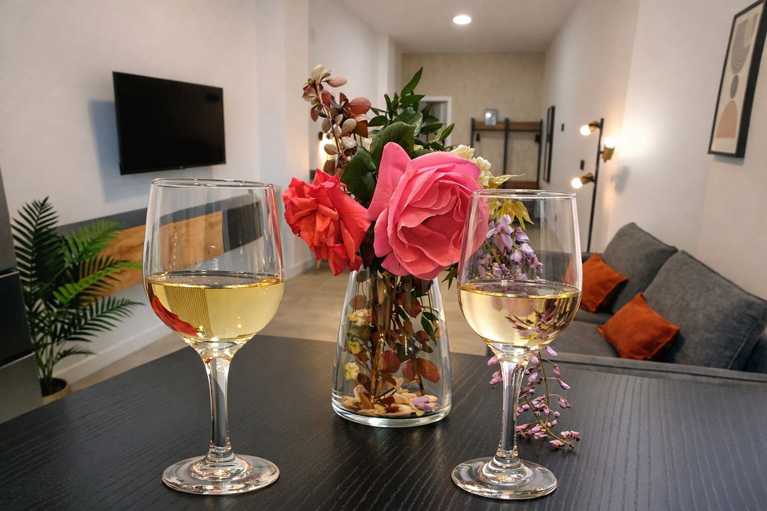 Una sala de estar moderna con una mesa de madera oscura, dos copas de vino blanco y un jarrón con flores rosadas y púrpuras.