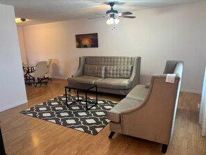 Un salon comprenant un grand canapé, une table basse, un tapis à motifs, un ventilateur de plafond et un tableau accroché au mur.