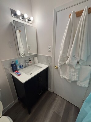 A bathroom with a white sink, dark cabinets, a mirror with lights, and a door with towels.