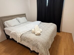 Un lit avec une couvre-lit à motifs et des oreillers, un jouet en peluche blanc posé sur le lit, et un rideau noir partiellement tiré.
