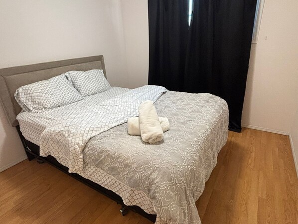 Una cama con una colcha y almohadas estampadas, un peluche blanco sobre la cama y una cortina negra entreabierta.