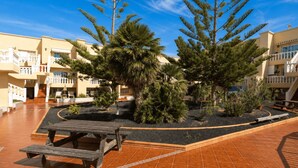Un patio con una mesa de picnic de madera, rodeado de plantas en macetas y árboles, con un edificio que tiene balcones y un piso de baldosas.