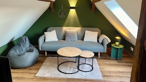 A loft bedroom with a sloped ceiling, a grey sofa, a bean bag, two small tables, and a wall-mounted lamp.