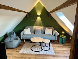 Une chambre à coucher aménagée dans un espace mansardé, avec un plafond en pente, un canapé gris, un fauteuil poire, deux petites tables et une lampe fixée au mur.