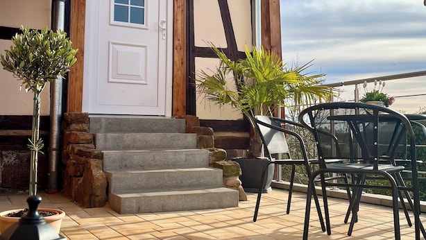A building with a white door, stone steps, a balcony with chairs, and potted plants.
