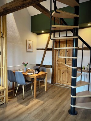 Een eethoek met een houten tafel en stoelen, een wenteltrap en uitzicht op de lucht via een dakraam.